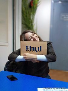 Gülümseyen bir kadın, önünde akıllı telefonuyla mavi bir masada otururken YENİ BİN YIL SÜRÜCÜ KURSU etiketli bir sertifika klasörüne sarılıyor.