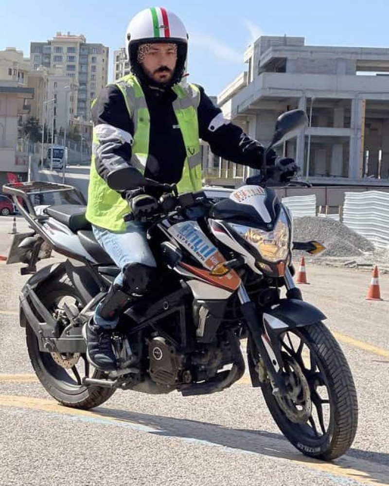 Kask ve yansıtıcı yelek giyen bir adam, arka planda yüksek binaların ve inşaatların göründüğü kentsel bir alanda siyah bir motosiklet sürüyor. Trafik konileri caddeyi kaplıyor.